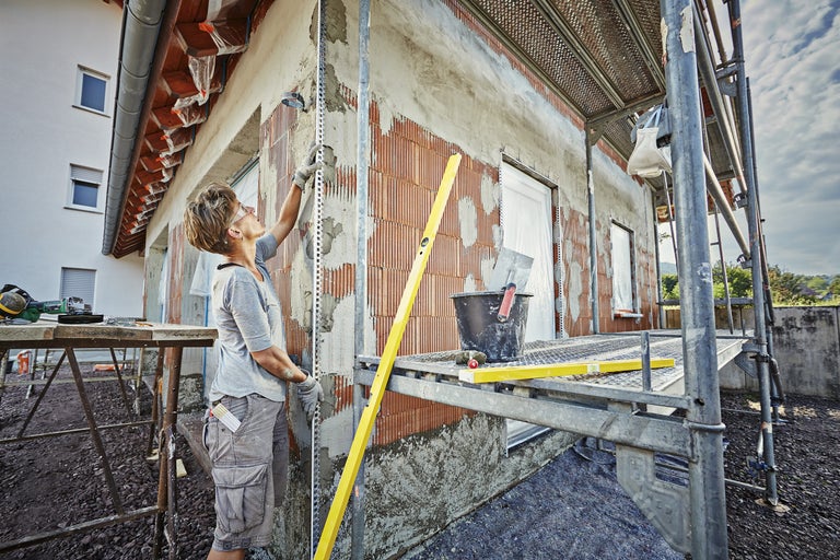 Artizan tencuind o fațadă de casă cu schelă, tencuială și unelte