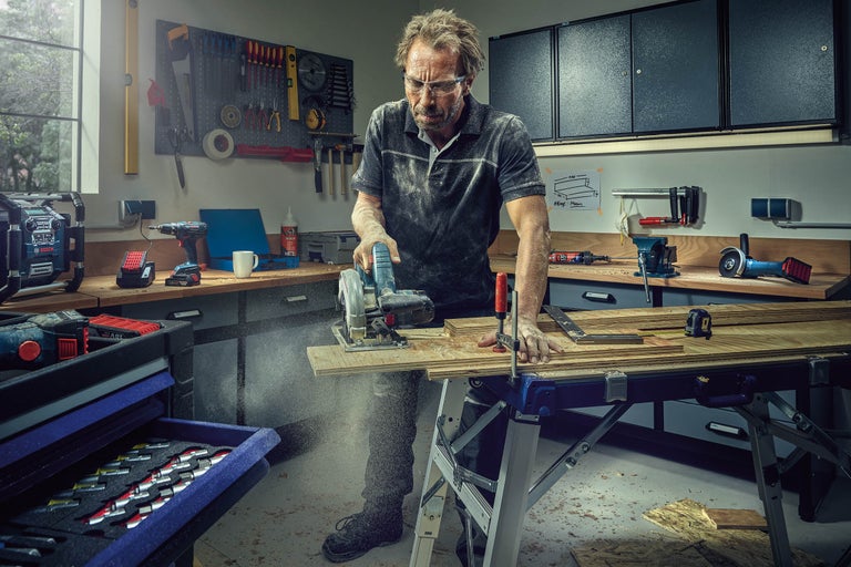 Un bărbat taie lemn cu un ferăstrău circular într-un atelier. Pe bancul de lucru se află bucăți de lemn, o menghină cu șurub și o ruletă.