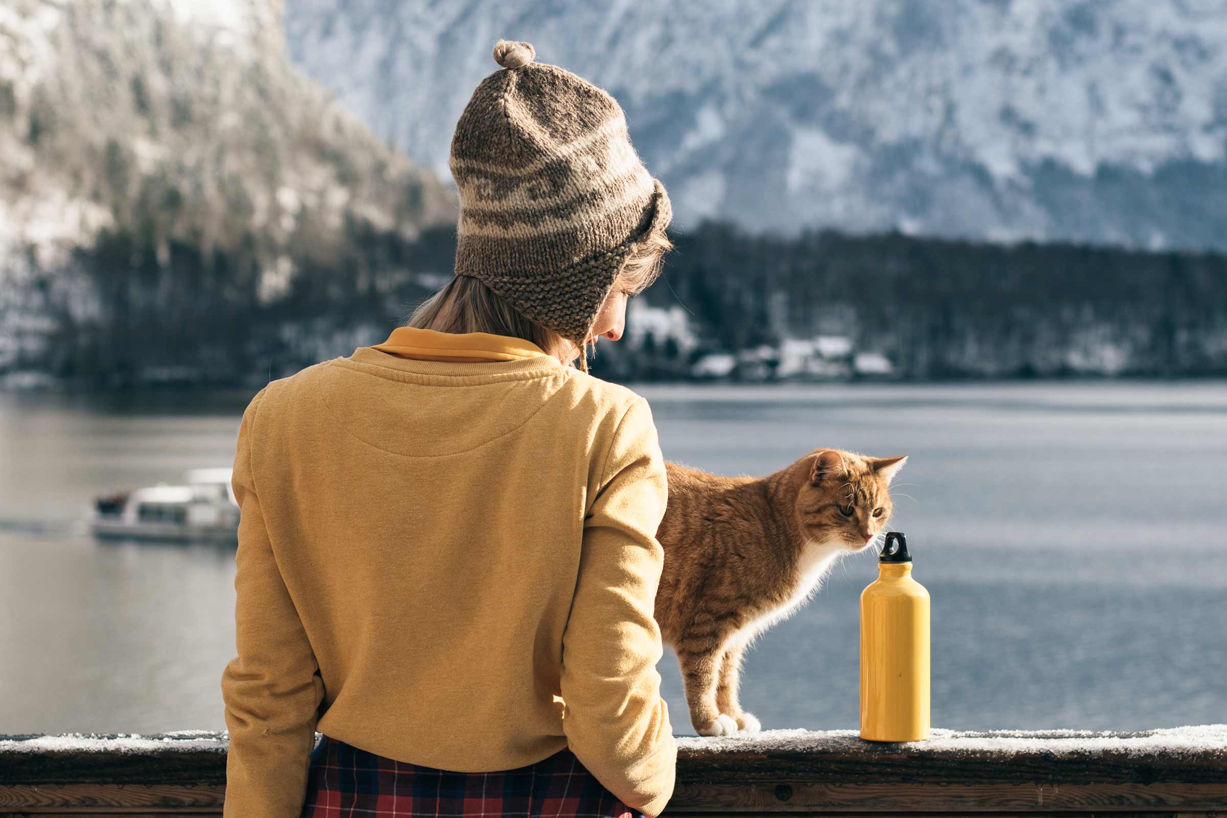 O femeie cu căciulă, o pisică și o sticlă galbenă de apă în fața unui lac montan