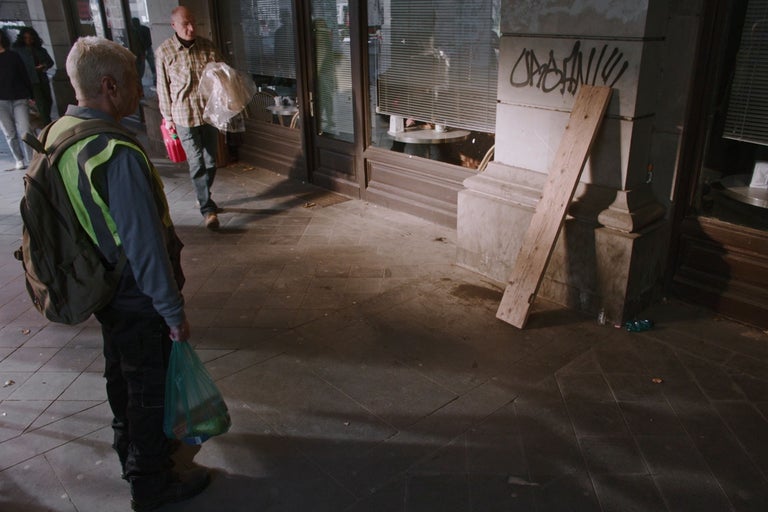 Scenă urbană de exterior cu o scândură de lemn pe o fațadă de piatră și un bărbat în vestă reflectorizantă în prim-plan.