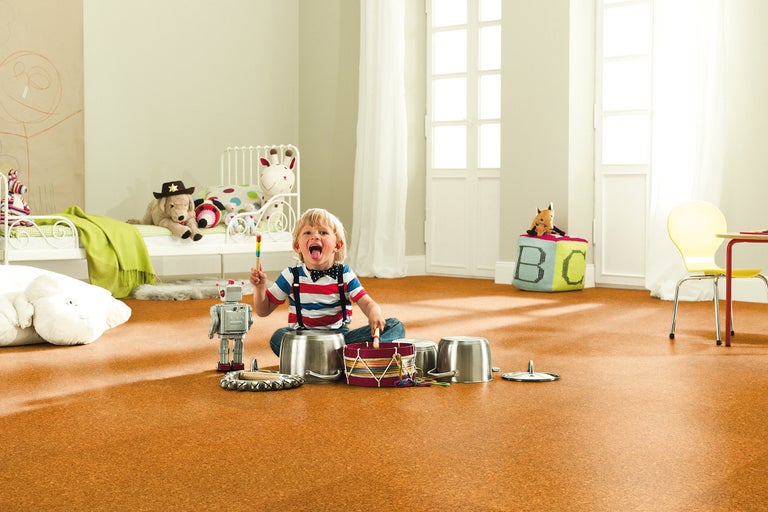 Cameră luminoasă pentru copii, cu podea de plută, unde un copil se joacă cu oale și un robot de jucărie. În fundal, se văd un pat și o zonă de relaxare.