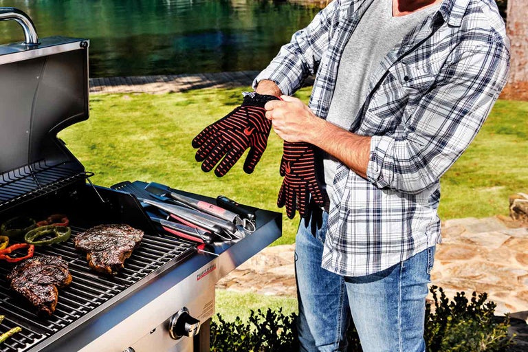 Un bărbat își pune mănuși de grătar pentru a se proteja de căldură în timp ce fripturi și legume sunt preparate pe grătar.