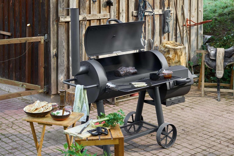 Scenă de grătar în aer liber cu un grătar afumătoare, mese și mâncăruri preparate.