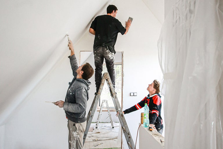 Trei meșteri renovează o cameră cu scări, vopsea și unelte de șlefuit.