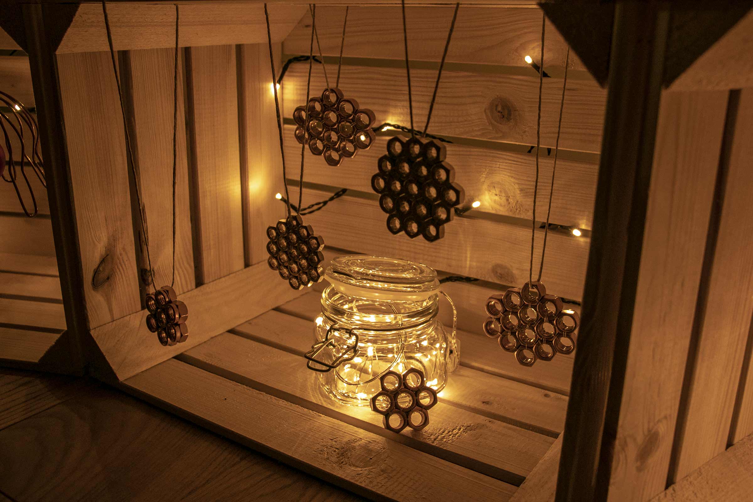 Scenă decorativă cu un borcan cu luminițe și pandantive din piulițe într-o ladă de lemn.