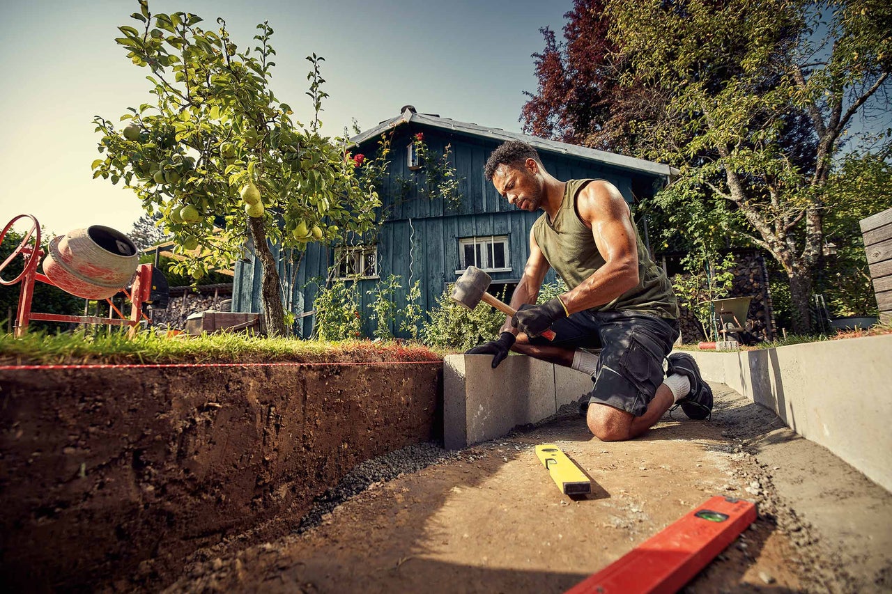 Un bărbat lucrează o piatră de pavaj cu un ciocan în grădină.