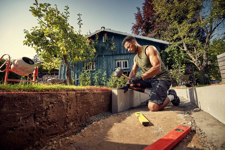 Un bărbat lucrează o piatră de pavaj cu un ciocan în grădină.