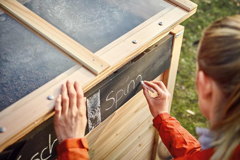 Prim-plan cu o seră rece din lemn, cu o persoană scriind cu creta „Spanac” pe o tablă.