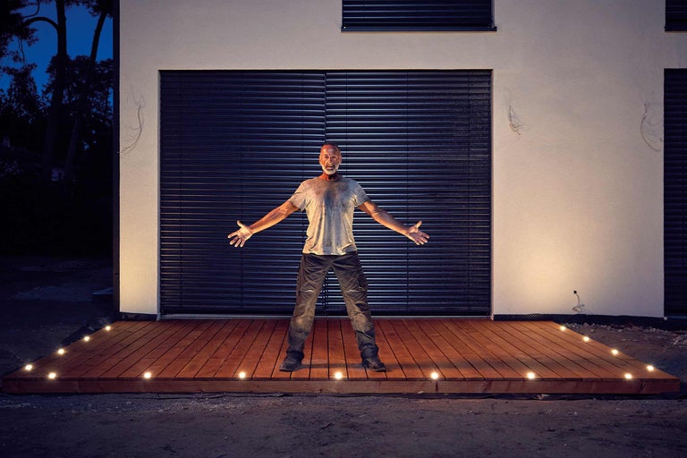 Un bărbat stă pe o terasă de lemn cu lumini integrate în fața unei case.