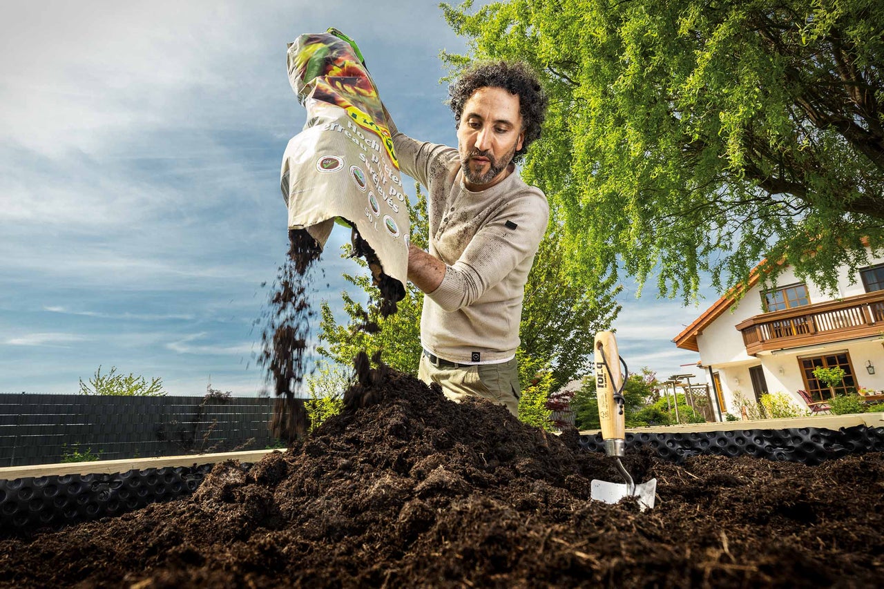 Un bărbat toarnă pământ de flori dintr-un sac într-o jardinieră înălțată în grădină.