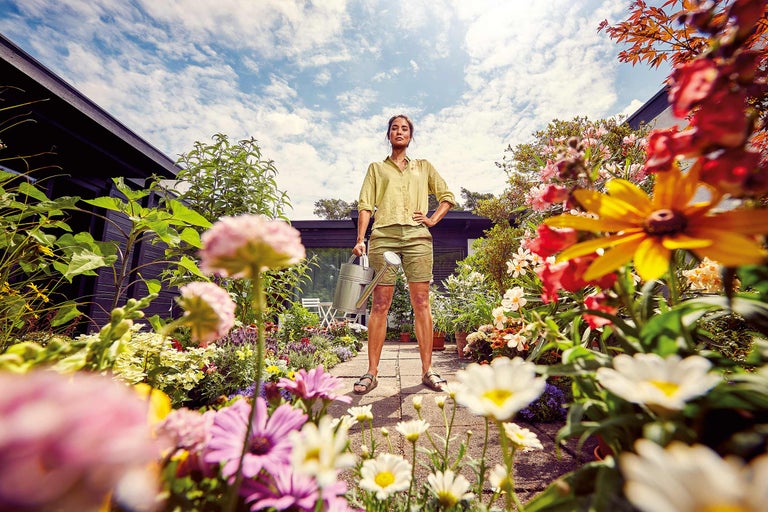 Femeie cu stropitoare stă într-o grădină plină de culoare.