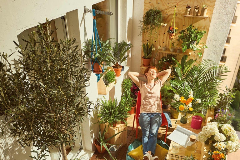 Femeie relaxându-se pe un balcon însorit, înconjurată de plante în ghivece și jardiniere suspendate.