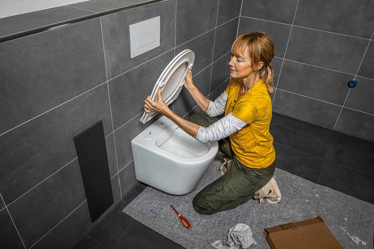 O femeie instalează un scaun de toaletă într-o baie cu gresie gri și un WC suspendat.