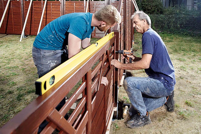 Doi bărbați construiesc un gard din lemn cu nivela și șurubelniță electrică.
