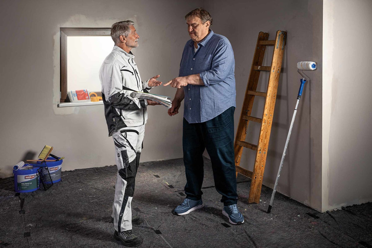 Doi oameni discută despre o renovare cu o scară, trafalet și găleți de vopsea într-o cameră.