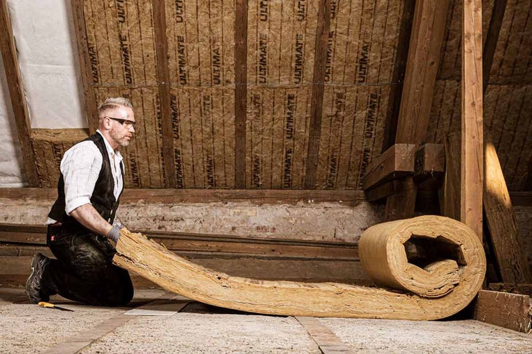 Un bărbat stă în genunchi într-un pod și derulează material izolant pentru a instala izolație termică.