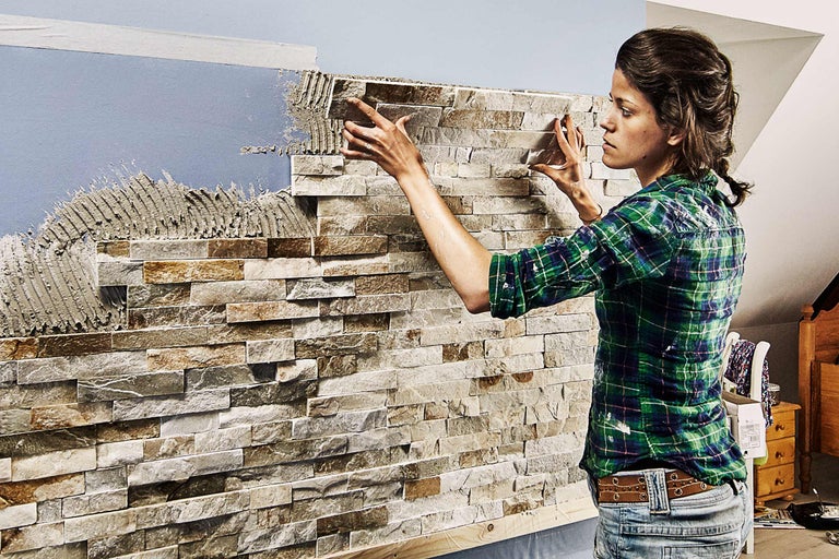 O femeie instalează panouri de piatră pe un perete folosind mortar adeziv.