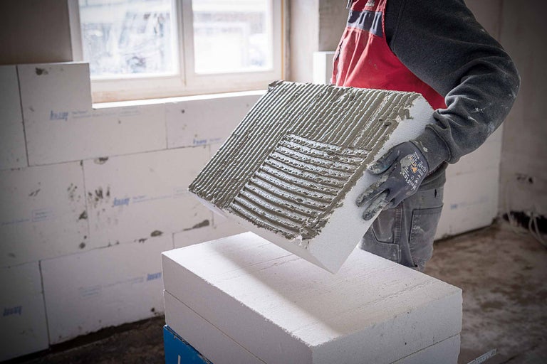 Un muncitor în construcții ține în mâini un bloc de beton celular cu adeziv aplicat pentru a construi un zid.