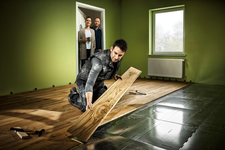 Un bărbat instalează podea laminată într-o cameră cu fereastră și calorifer. În fundal, două persoane stau într-o ușă deschisă.