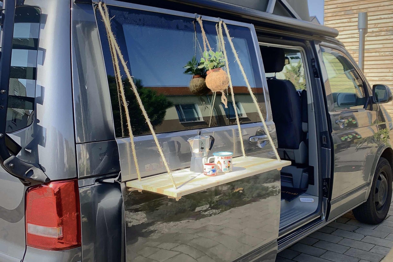Furgonetă de camping cu masă suspendată atașată la ușa din spate, cu băuturi și plantă
