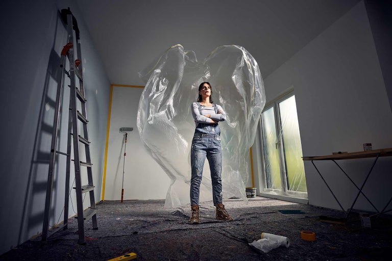 Femeie stând cu brațele încrucișate într-o cameră care necesită renovare cu o scară și o folie de acoperire