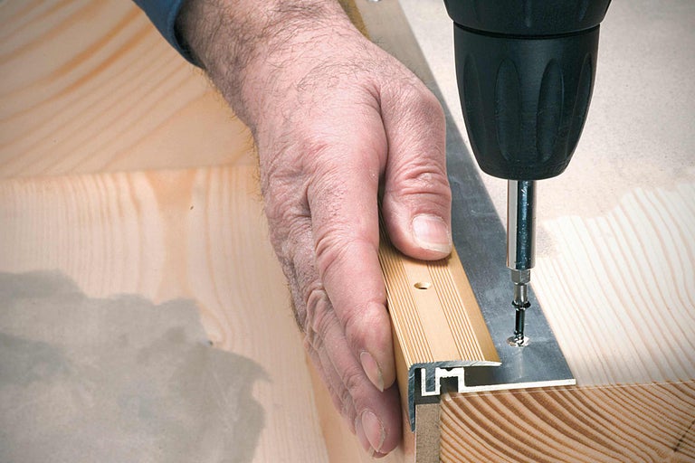 Mână fixează profilul de capăt pe podeaua de lemn cu șurub și șurubelniță cu acumulator