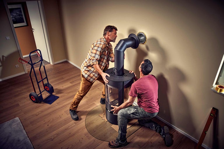 Doi meseriași instalează o sobă de lemne într-o sufragerie cu podea laminată și un cărucior.