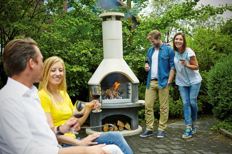 Scenă cu grătar de grădină pe care arde lemn și patru persoane care se relaxează în grădină cu pahare de vin.