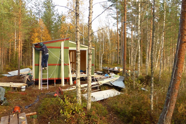 În pădure se construiește o casă de lemn. O persoană lucrează pe o scară la fațada casei.