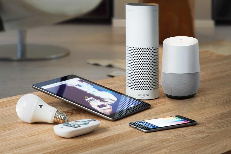 Scenă cu dispozitive inteligente pentru casă: tabletă, smartphone, difuzoare de la Amazon și Google, lampă LED și telecomandă pe o masă de lemn.