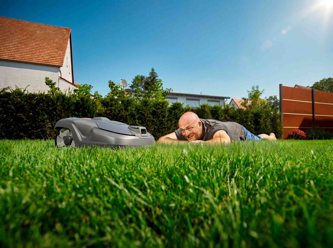 Un bărbat stă întins pe gazon lângă un robot de tuns iarba într-o grădină luminoasă și însorită.