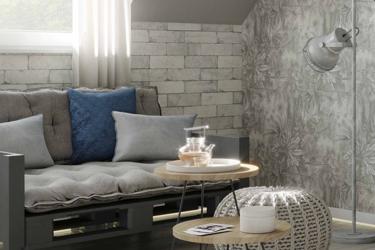 Cameră de zi cu canapea din paleți, perne, măsuțe laterale, taburet tricotat și lampă de podea în fața unui tapet cu model