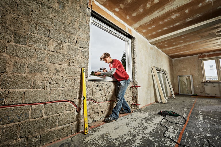 Un bărbat montează o fereastră cu o șurubelniță cu acumulator într-o clădire în construcție. O nivelă cu bulă de aer este sprijinită de perete.