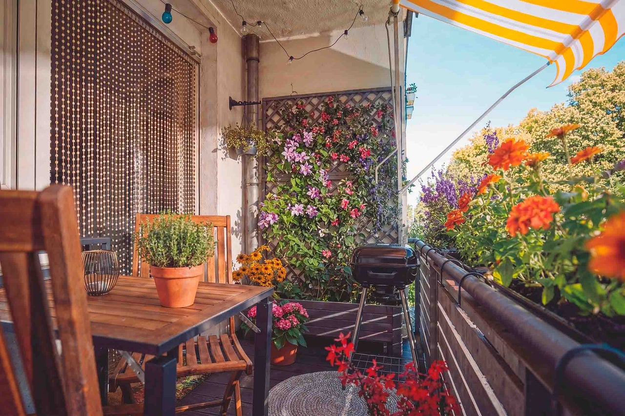 Balcon confortabil cu mobilier din lemn, grătar cu cărbuni, flori în ghivece, spalier cu plante cățărătoare și marchiză cu dungi galbene și albe.