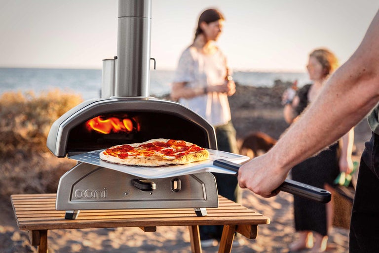 Cuptor de pizza Ooni cu paletă de pizza pe o masă de lemn, cu oameni pe plajă în fundal.