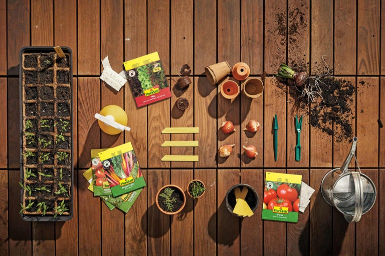 Scenă de grădină cu tavă de însămânțare, plicuri de semințe FloraSelf, ghivece de flori și unelte de grădinărit pe podea de lemn.