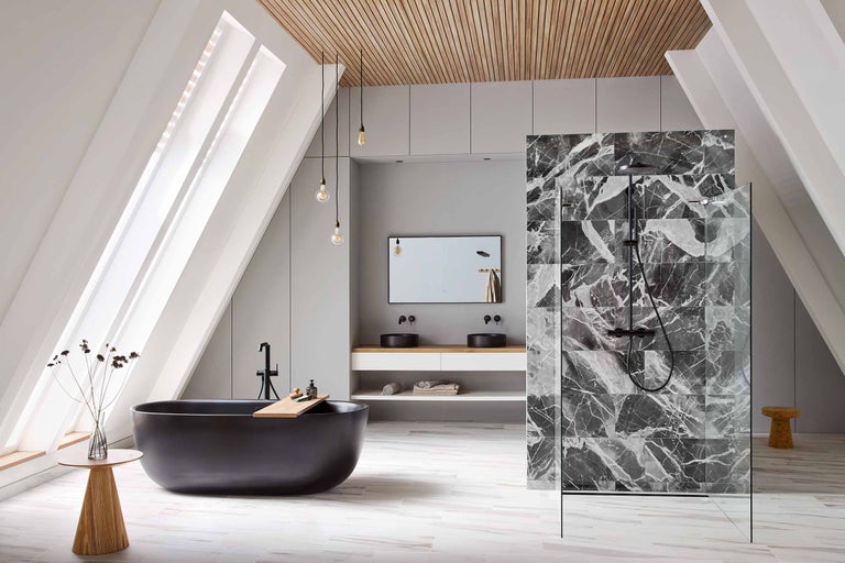 Baie luminoasă cu pereți înclinați, cadă neagră, dulap de lavoar cu două chiuvete, zonă de duș cu plăci de marmură și elemente din lemn