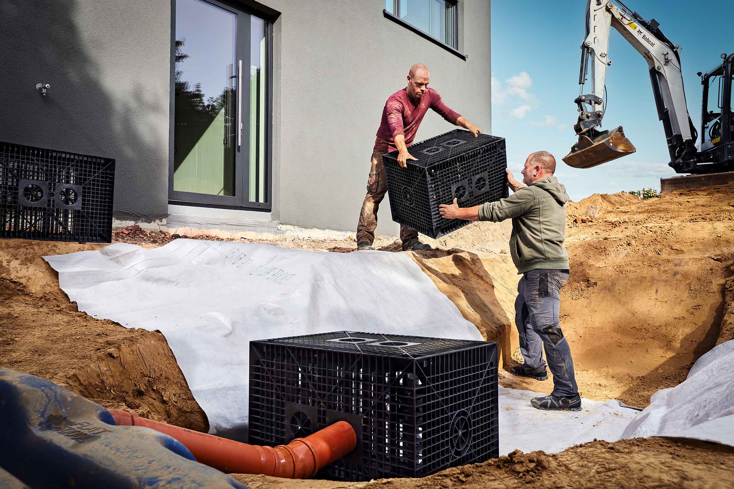 Doi bărbați instalează blocuri de infiltrare pentru infiltrarea apei pluviale într-o groapă de construcție, căptușită cu un material geotextil alb.