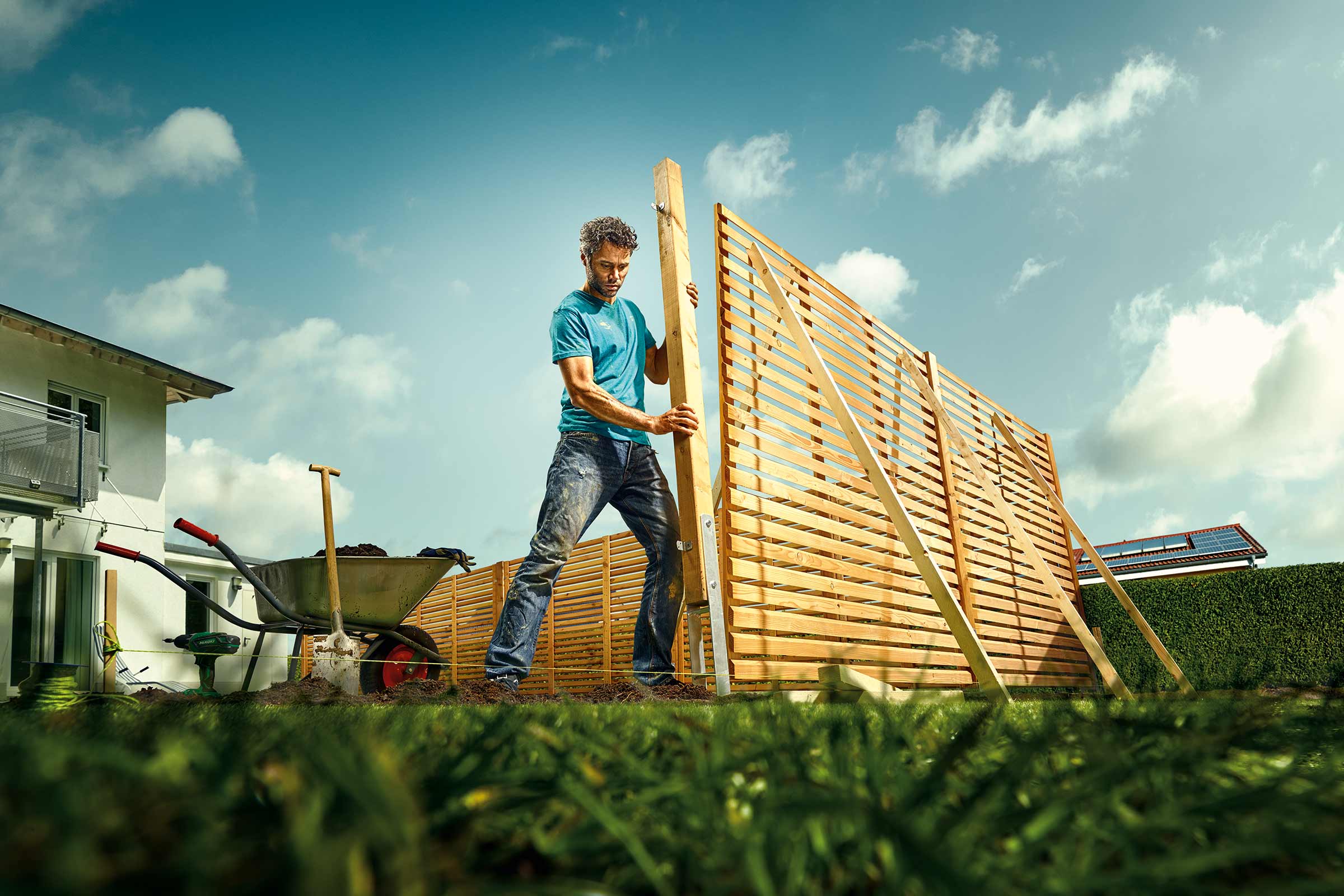 Bărbat construind un gard de lemn în grădină. Roabă și unelte vizibile în imagine.