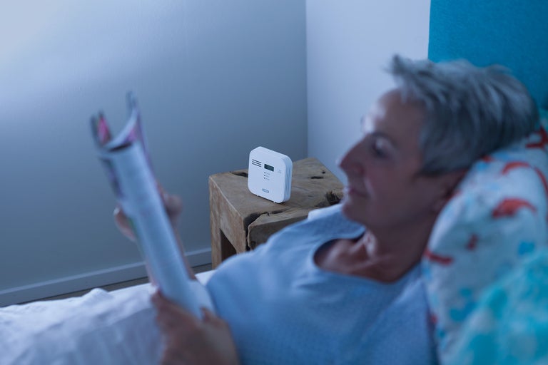 O persoană stă întinsă în pat și citește, cu un detector de monoxid de carbon pe noptieră.