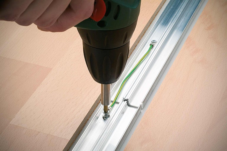 O persoană înșurubează o șină de profil din aluminiu pe o podea de lemn cu o șurubelniță electrică.