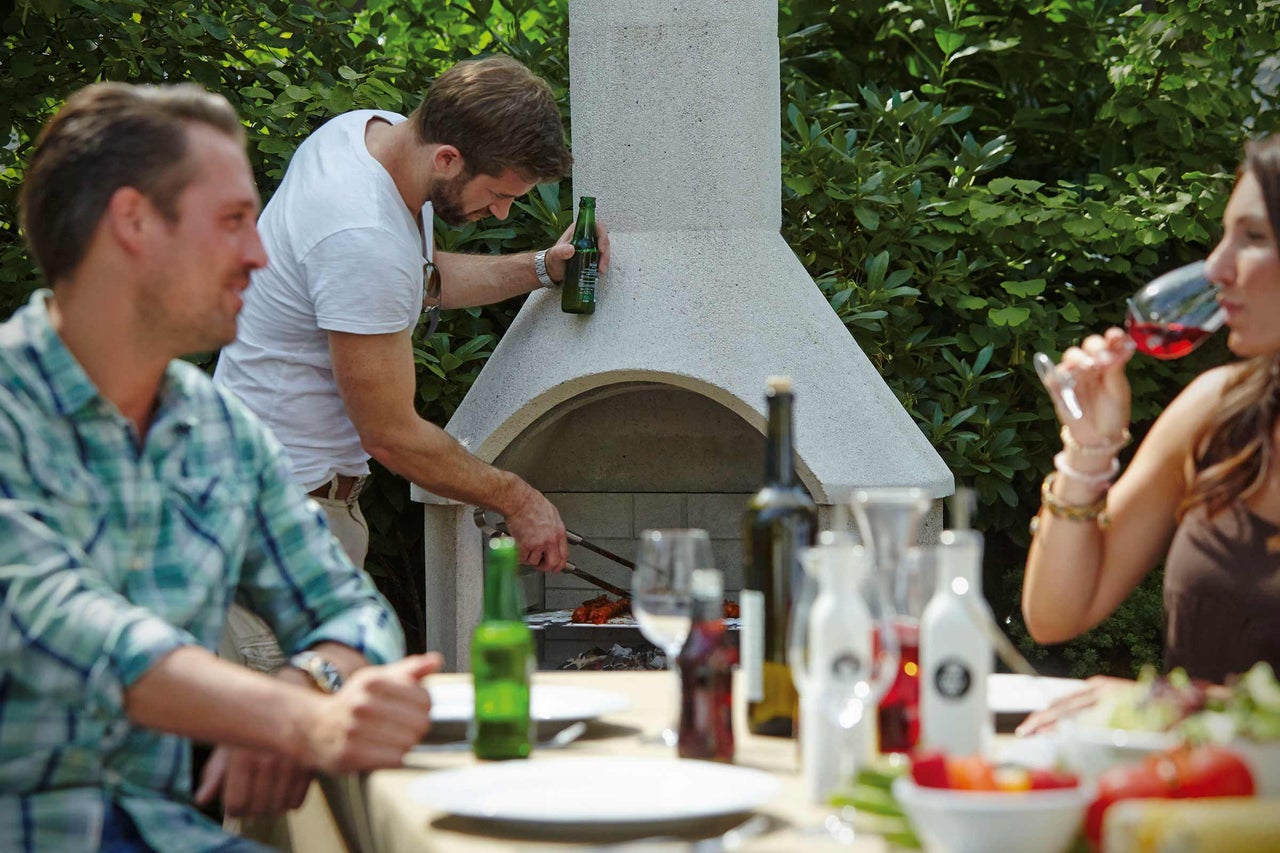 Persoane care fac grătar în grădină cu un grătar de grădină din piatră și savurează mâncăruri și băuturi la o masă.