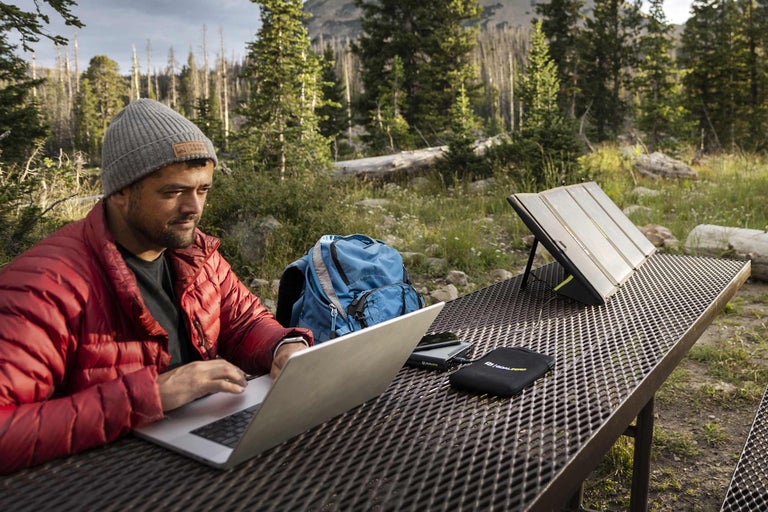 Un bărbat lucrează la un laptop cu un panou solar portabil pe o masă în aer liber