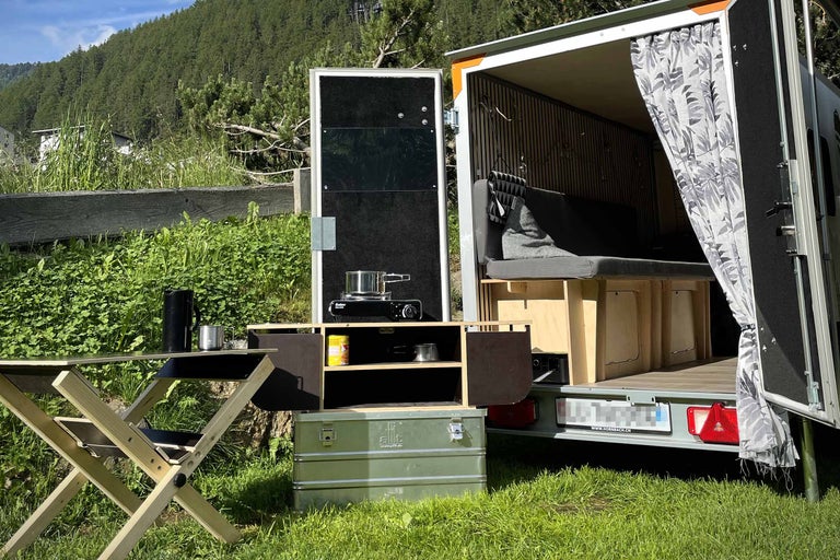 Rulotă de camping cu bucătărie, masă și spațiu de depozitare în aer liber.