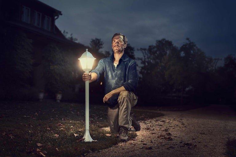 Bărbat îngenuncheat lângă o lampă de exterior aprinsă în grădină, noaptea.