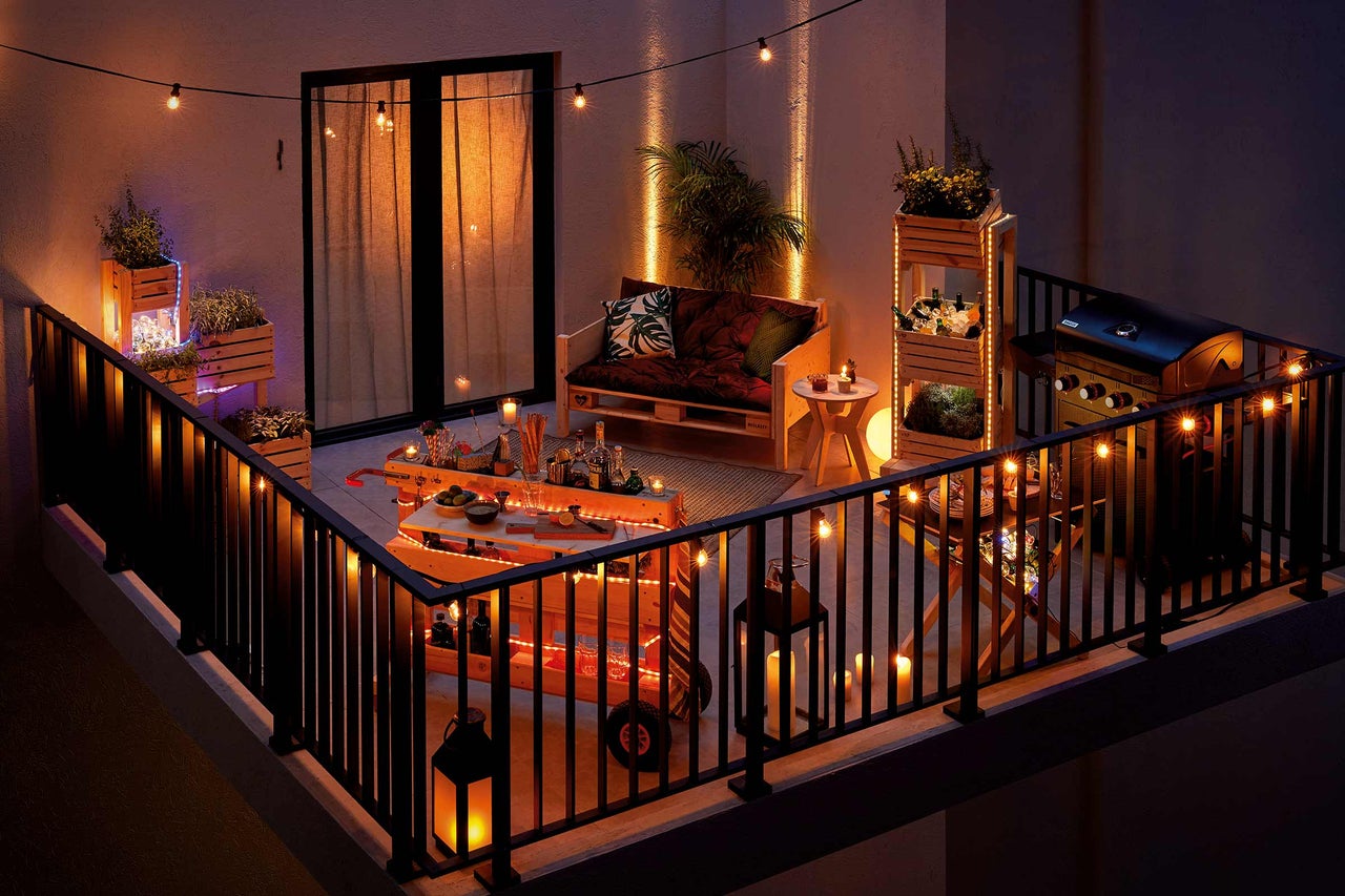 Scenă atmosferică de balcon cu grătar, cărucior de bar și mobilier din paleți, decorată cu lanțuri luminoase și jardiniere