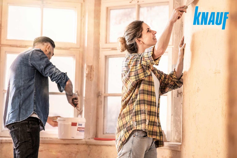 Două persoane gletuiesc un perete cu chit Knauf într-o cameră luminoasă.