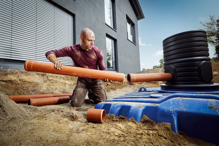 Un bărbat instalează conducte de drenaj pe un rezervor de infiltrare albastru în fața unei case.