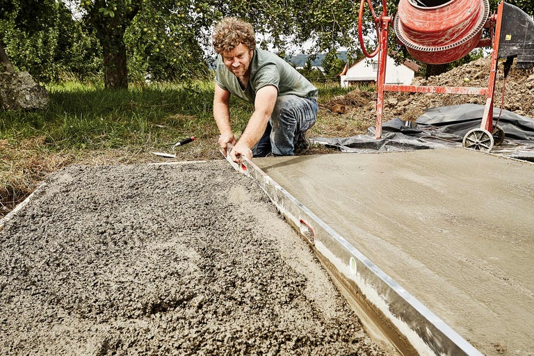 Bărbat nivelează betonul cu o nivelă cu bulă de aer, cu un mixer de beton în fundal.