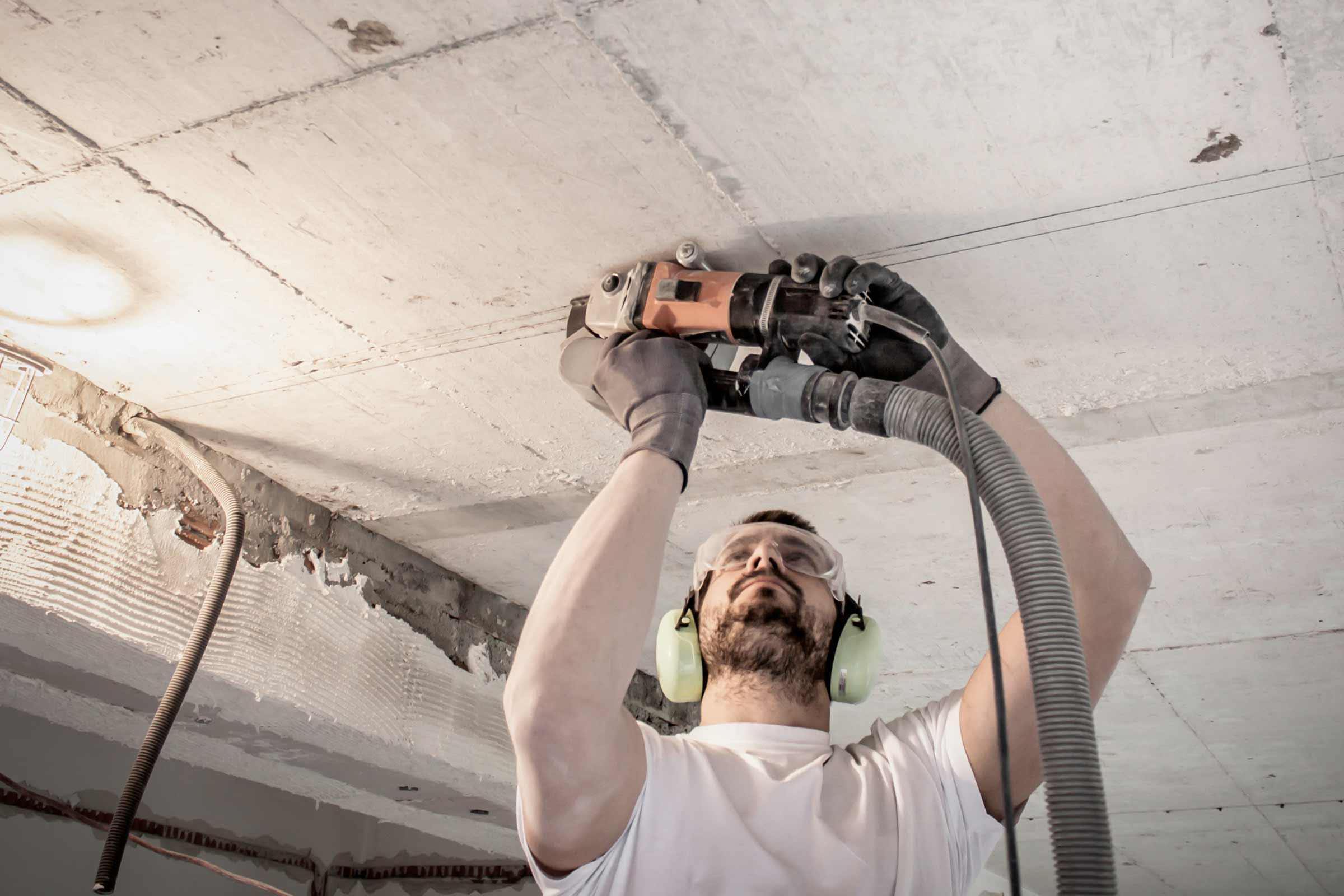 Un bărbat șlefuiește un tavan de beton cu o mașină de șlefuit beton, purtând ochelari de protecție și protecție auditivă